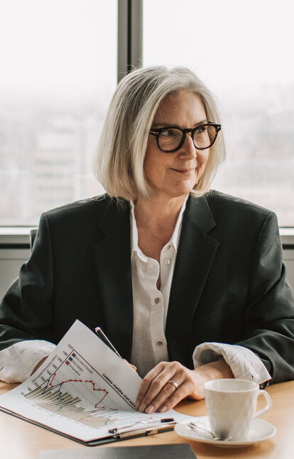 A grey-haired business owner in a dark blazer smiles at a desk, a coffee and financial charts in front of her