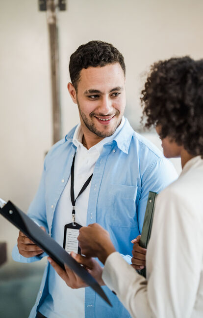 Two smiling professionals in conversation, one holding a clipboard and a lanyard badge